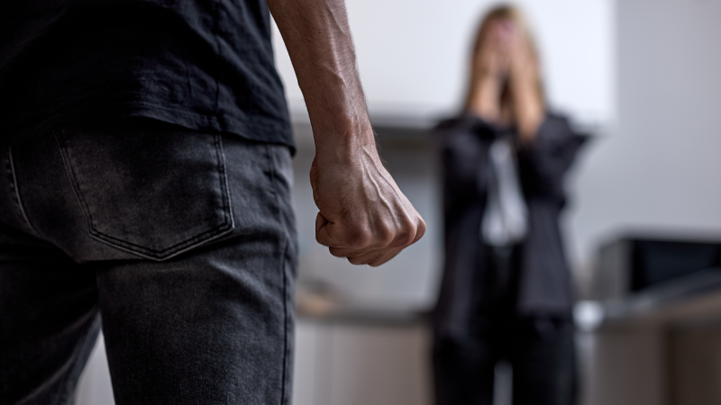 Aggressive Angry Husband Going To Beat Unhappy Crying Wife Standing In Background. Family Conflicts And Quarrels, Married Life With Abuser. Domestic Violence Concept. Focus On Male Fist