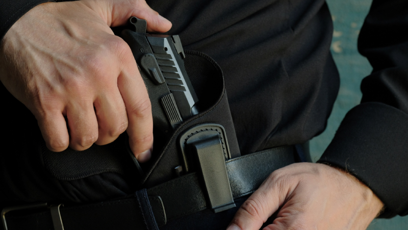 A man in black clothes takes out a black pistol from the holster, which is located on the waist belt of his trousers. Concept: security, possession of firearms.