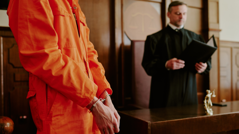 Caucasian young adult man in orange jumpsuit standing in handcuffs before judge in courtroom, judge holding folder and standing behind bench, legal proceedings in progress
